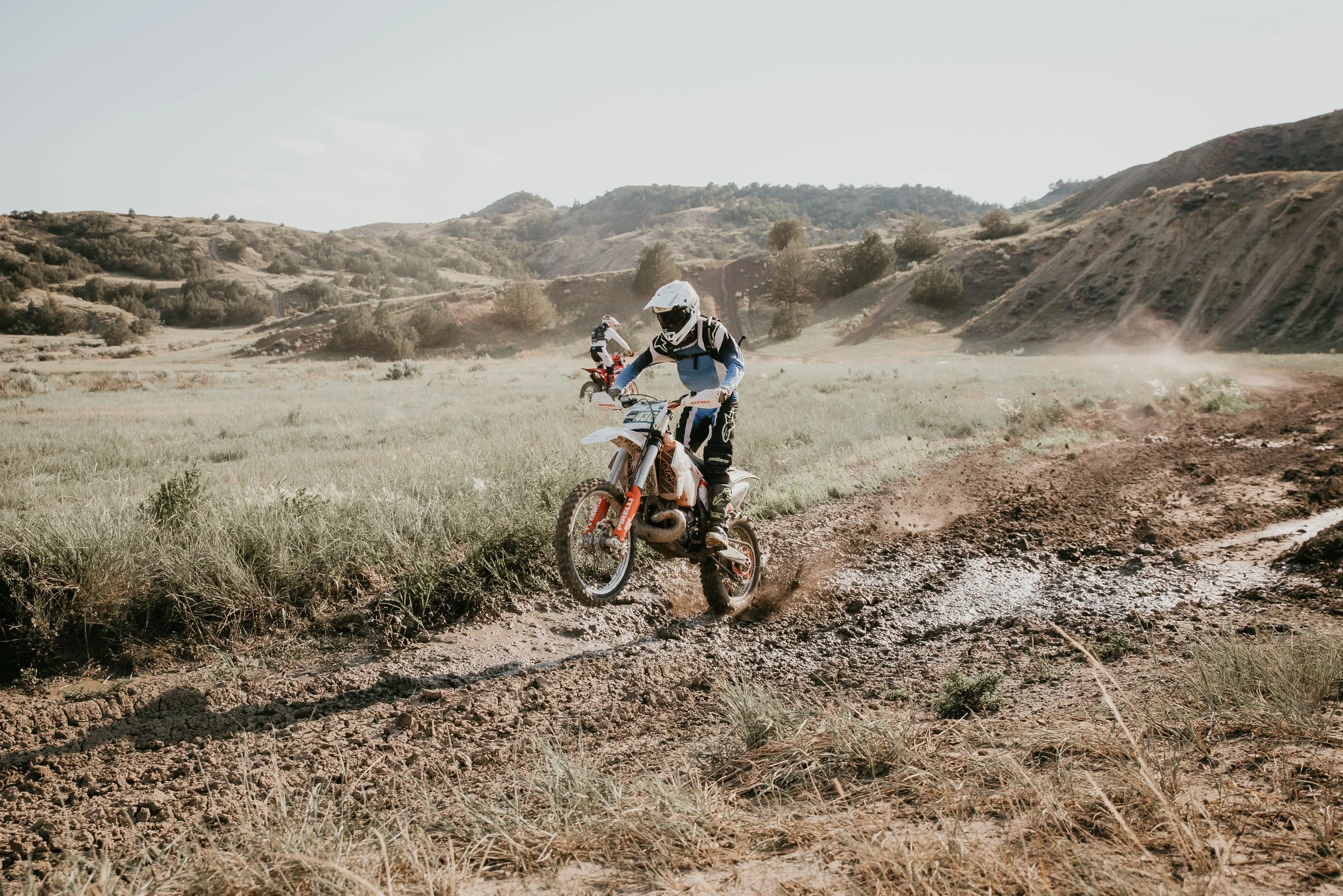 Two motocross riders on dirt bikes riding through a dusty off-road trail in a hilly, semi-arid landscape with sparse vegetation.