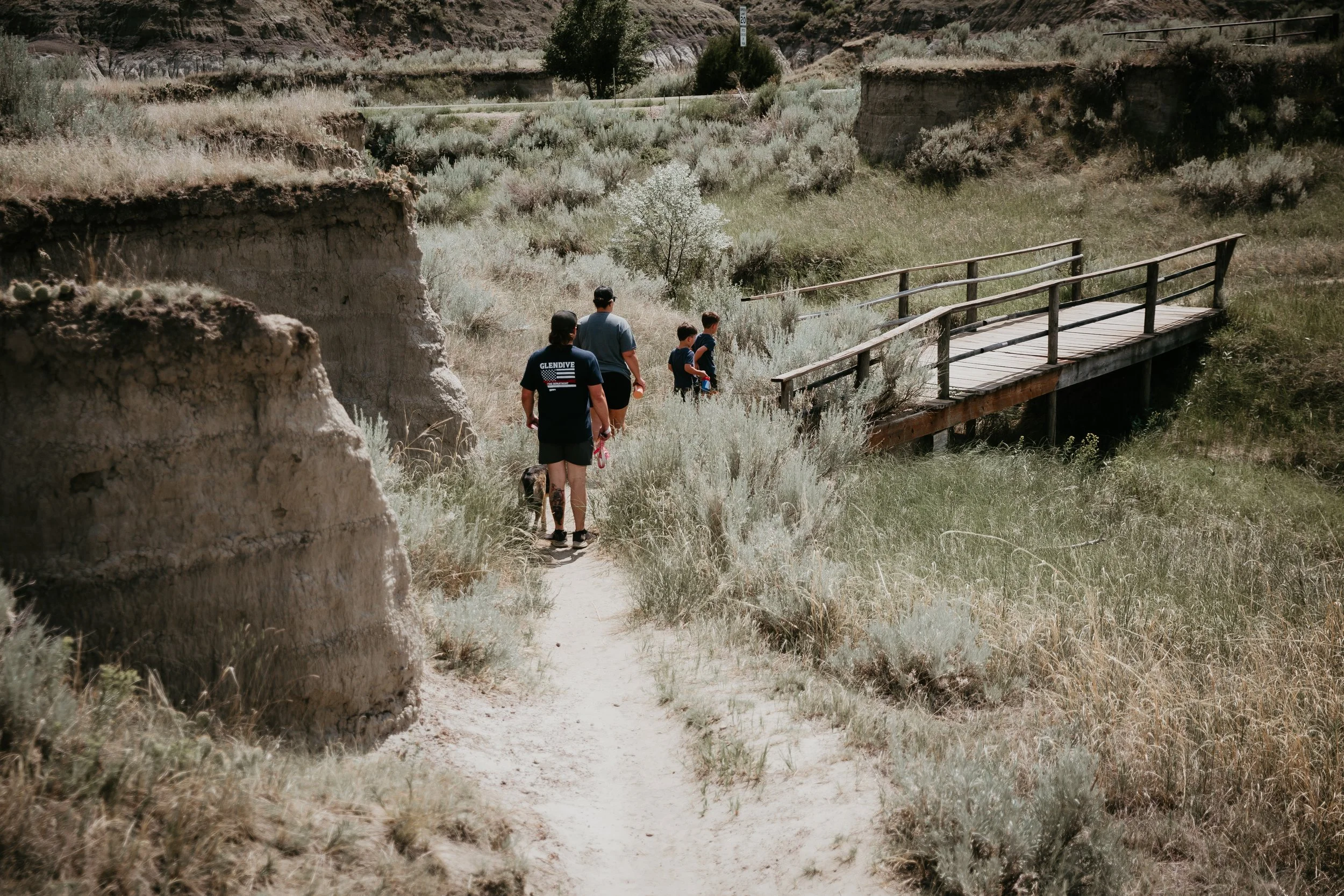 A family of five with a dog walking on a trail through a dry, desert landscape with sparse vegetation, cliffs, and a wooden bridge in the background.