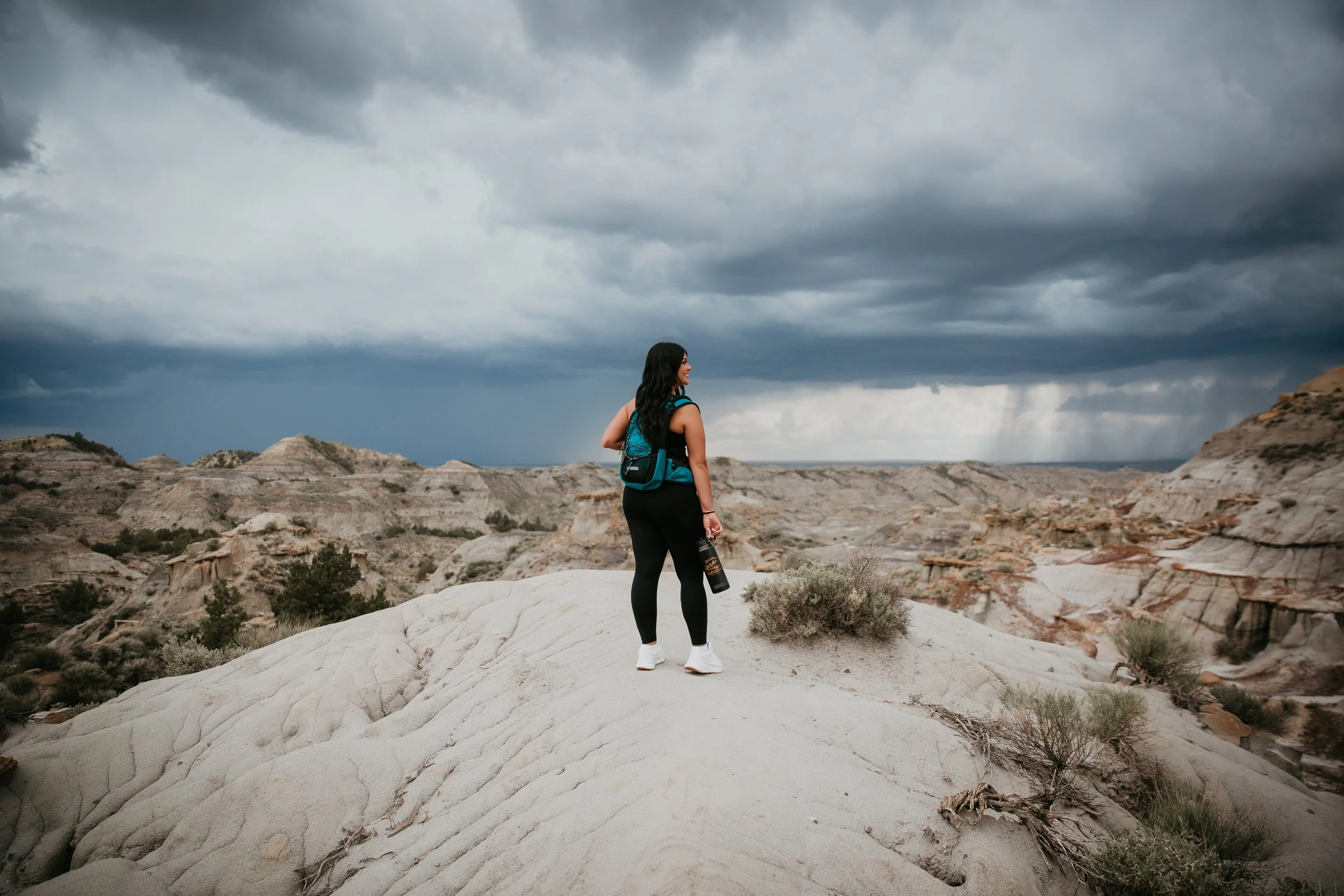 A woman with long dark hair, wearing a blue backpack, black leggings, white shoes, stands on a white rocky landscape beneath dark, stormy clouds. She is holding a black water bottle and looking away from the camera toward the horizon.