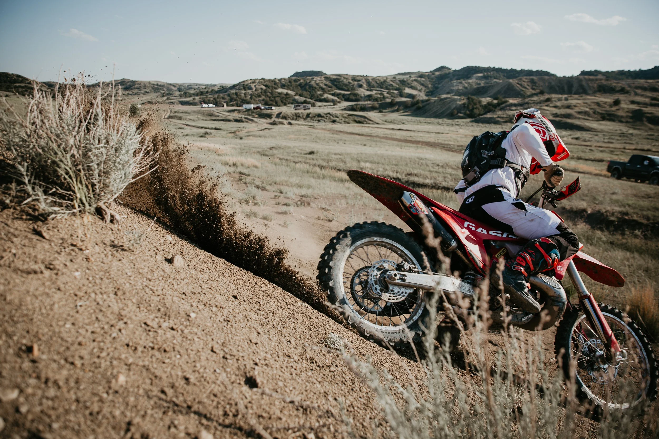 A motocross rider in white gear riding a red dirt bike on a dusty trail in a desert landscape with hills and sparse vegetation.