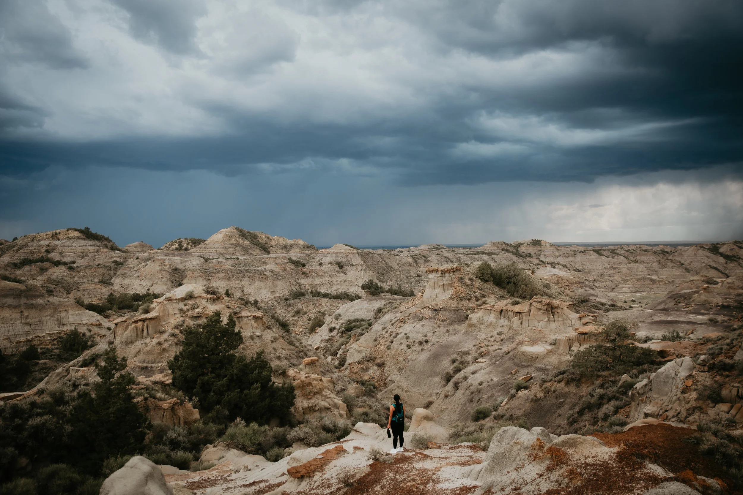 A lone hiker with a backpack walking on rocky terrain through a desert landscape with eroded rock formations and sparse vegetation, under a dark, cloudy sky.
