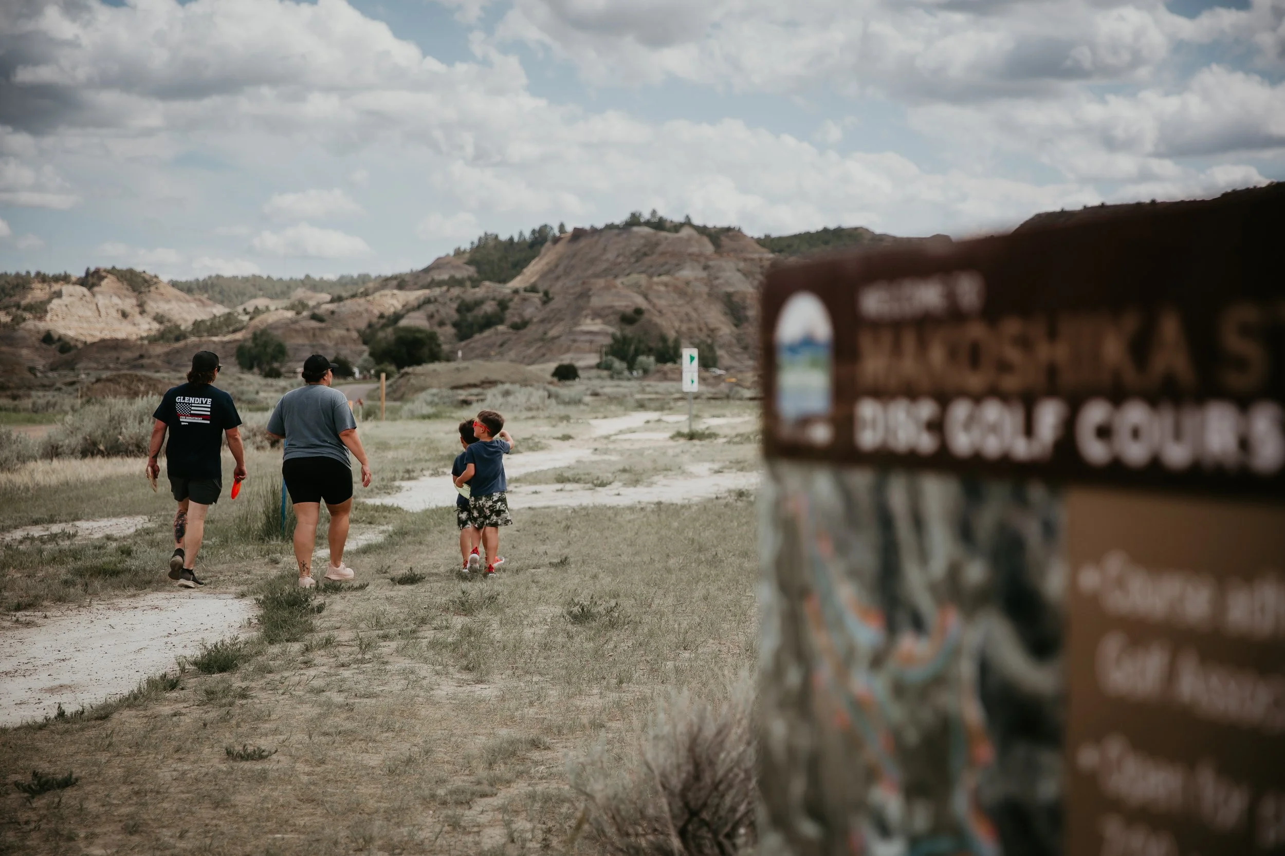 A group of four people, including two children, walking towards the Makoshika State Park Disc Golf Course under a cloudy sky. 