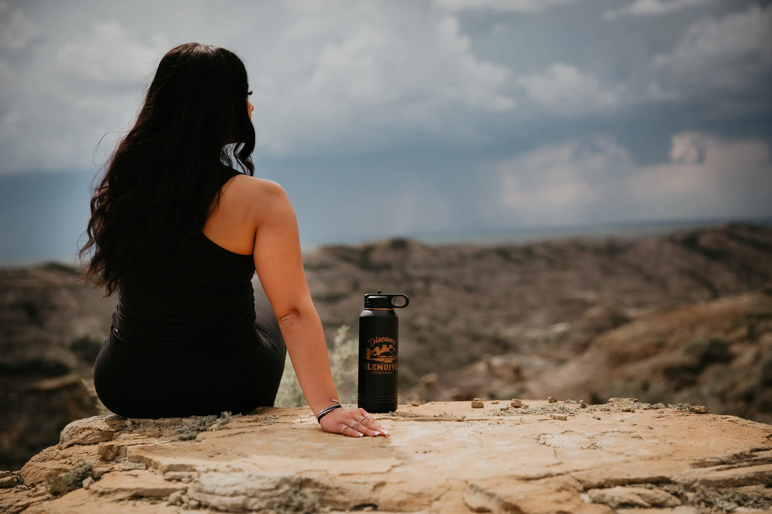 A woman with long dark hair sitting on a rock in a desert landscape, looking away from the camera at cloudy skies, with a water bottle branded 'Discover Glendive Montana' beside her.
