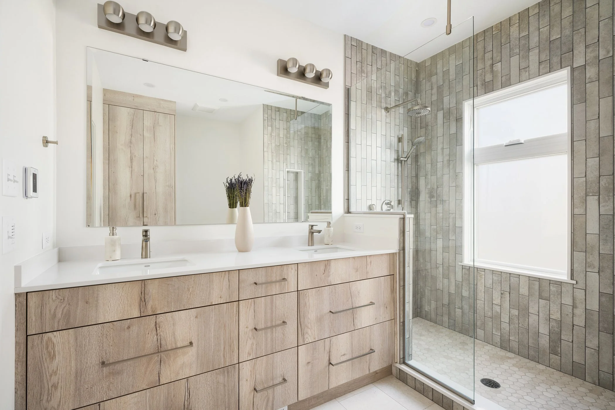 Modern St. Paul bathroom with doorless walk-in shower featuring vertical gray tile, frameless glass, dual vanity with wood cabinets, and bright natural lighting.