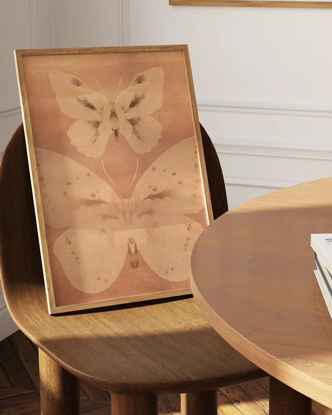 A framed print of a butterfly with symmetrical wings, placed on a wooden chair next to a round wooden table with some books on it, inside a room with white walls.