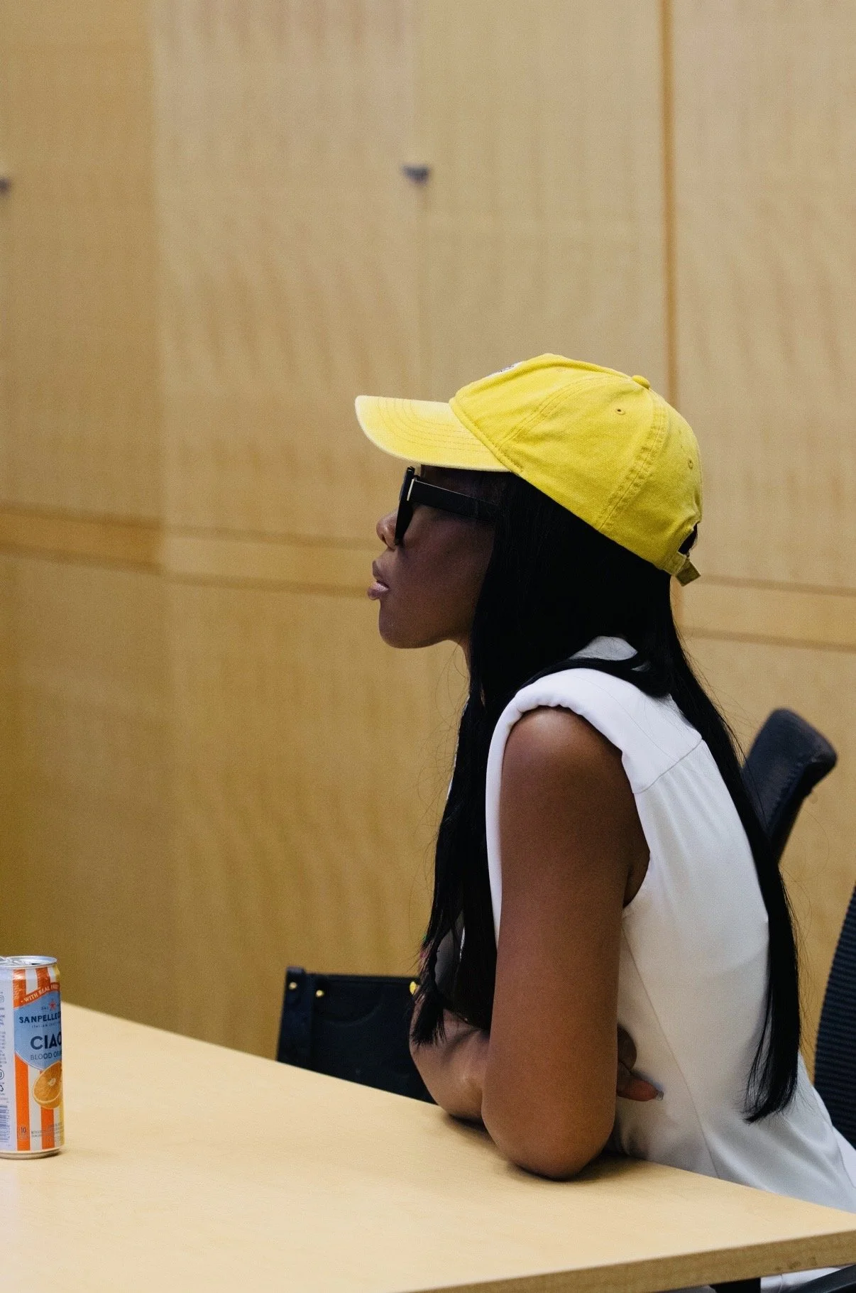 Side profile of a woman wearing a yellow baseball cap, black sunglasses, and a sleeveless white top, sitting at a table in a room with wooden walls.