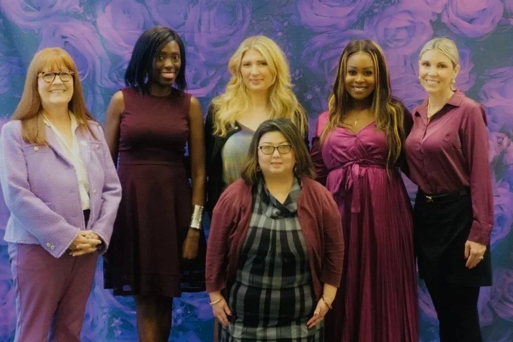Six women standing in front of a purple floral backdrop, smiling at the camera.