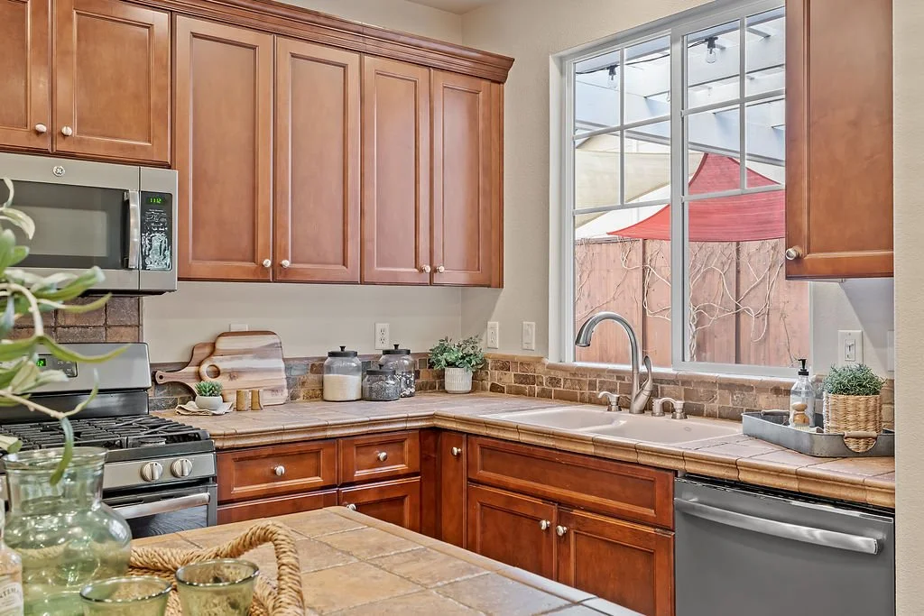 Kitchen with wooden cabinets, stainless steel microwave and oven, large window above the sink, brick backsplash, and various decorative items on the countertop.