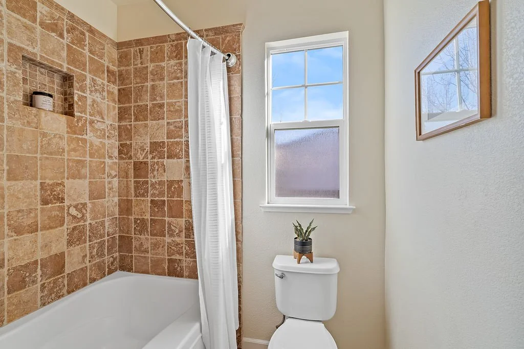 Bathroom with a bathtub and shower area covered by a white shower curtain, tan tiled walls, small niche in tiles, toilet with a potted plant on top, window with divided panes letting in natural light, and framed picture on the wall.