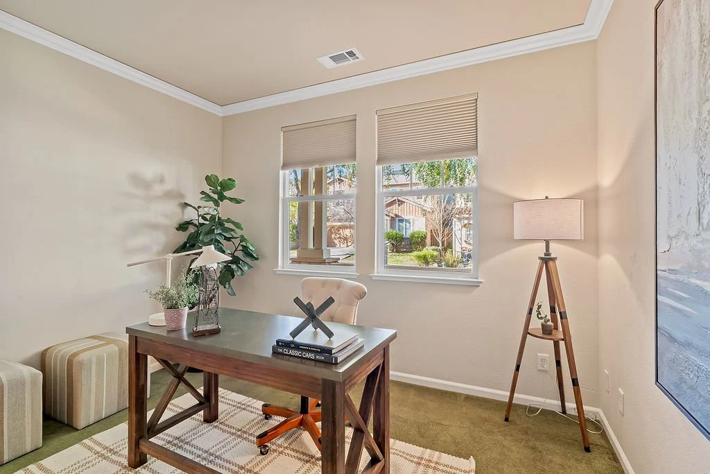 A home office with beige walls, a wooden desk, a beige chair, a large potted plant, two beige upholstered stools, a desk lamp, a floor lamp with a beige shade, a window with blinds showing a neighborhood view, and a large abstract painting on the wal