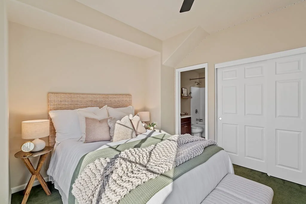 A bedroom with a bed that has multiple pillows and a textured throw blanket. There are two circular bedside lamps on wooden side tables, and a closet with sliding doors. An open doorway reveals a small bathroom with a toilet and shelf.