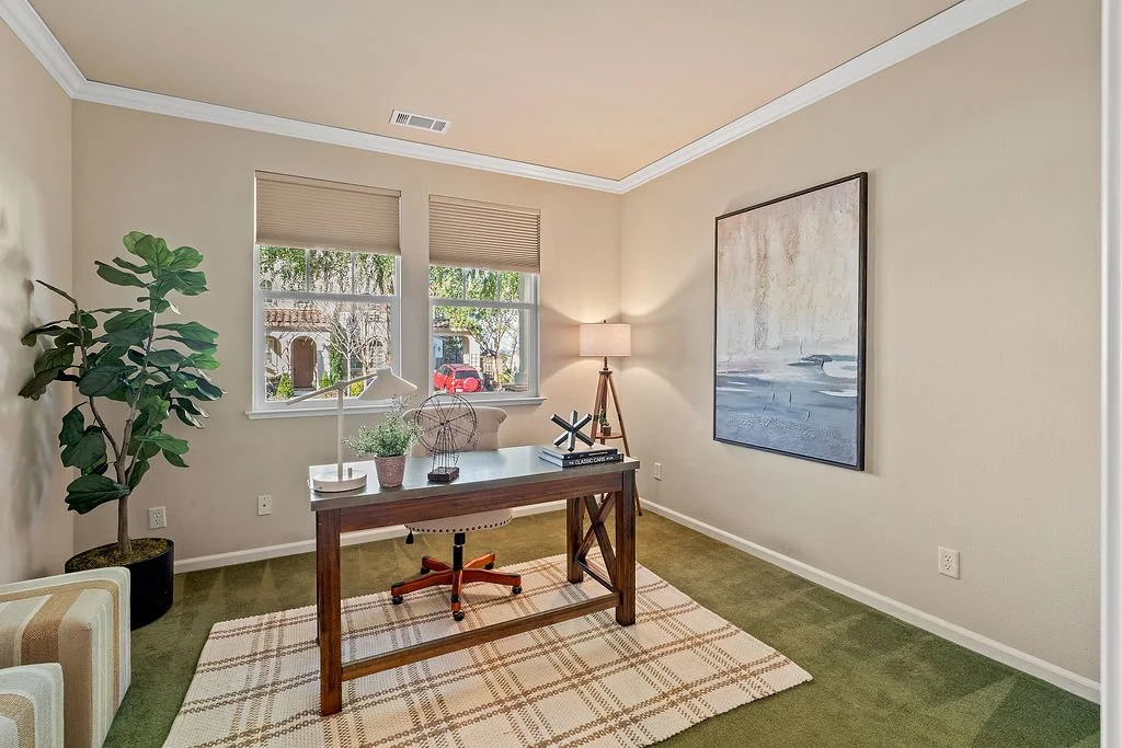 Home office with a desk, office chair, potted plant, lamp, abstract artwork, and large windows with blinds, carpeted floor, and neutral-colored walls.