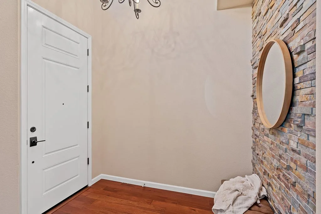 An entryway with a white door, beige walls, a stone accent wall with a circular mirror, a chandelier overhead, and a pile of cloth on the floor.