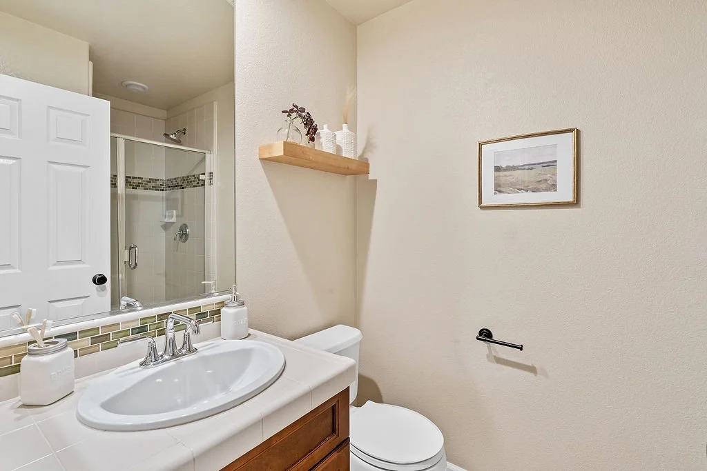 Bathroom with a white sink and cabinet, a mirror, a toilet, and a shower with glass door. The wall has a framed picture and a floating shelf with decorative items.