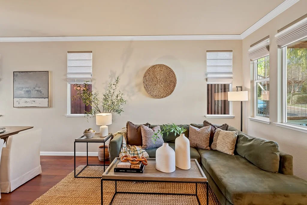 Living room with beige walls, brown wood flooring, green sectional sofa with pillows, two white decorative vases on coffee table, small side table with a lamp and bowl, artwork and windows with blinds, and a floor lamp.