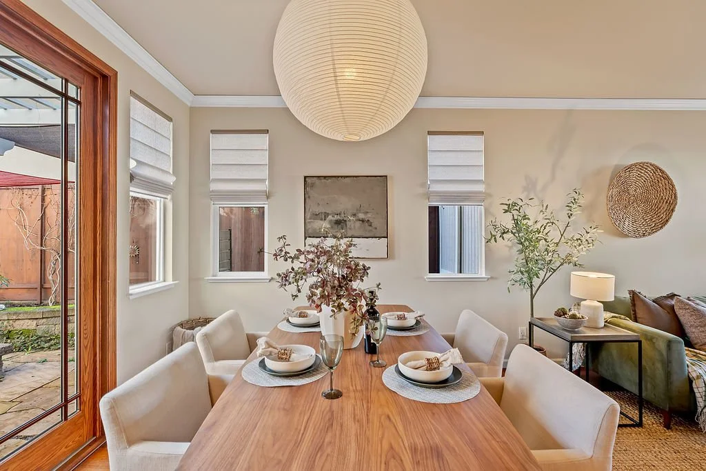 Dining room with a wooden table set for six, decorated with a large floral centerpiece, surrounded by beige upholstered chairs, and featuring a paper lantern ceiling light, three small windows with white blinds, and a sliding glass door leading to an