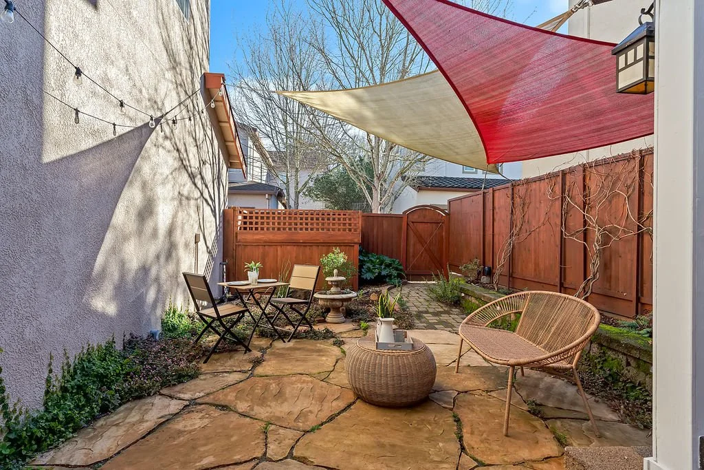 A cozy backyard patio with stone paving, outdoor seating including a round wicker table, a curved wicker chair, and two folding chairs around a small round table. There are string lights overhead, a birdbath, potted plants, a privacy fence, and fabri