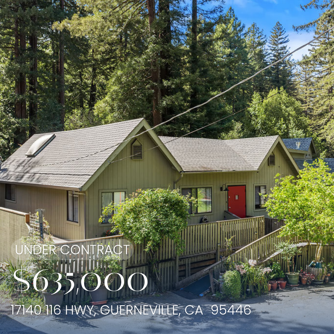 Photo of a house in a wooded area with a fenced yard, trees, and potted plants. The house features a gray exterior, red front door, and a sloped roof with solar panels and a skylight.