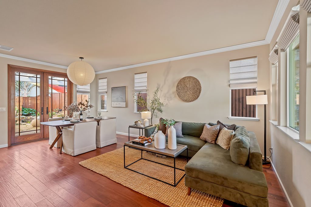 Living room with green sectional sofa, coffee table with white vases, and artwork on wall. Adjacent dining area with table and chairs, large window, and sliding glass door opening to outdoor space.