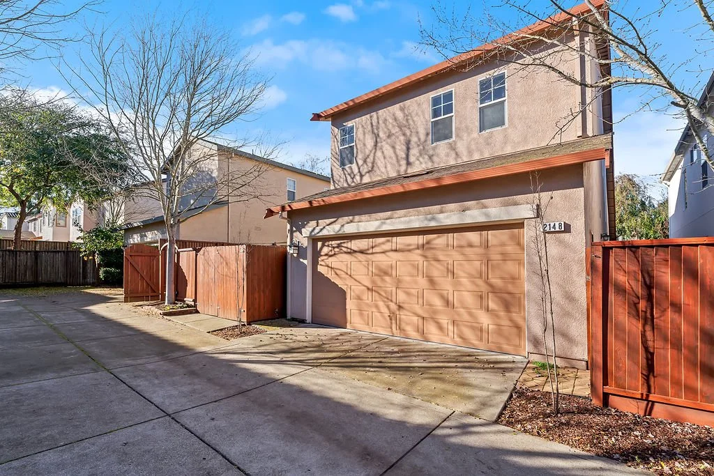 A two-story house with a closed garage door, a red fence, two small trees, and a concrete driveway under a blue sky with some clouds.