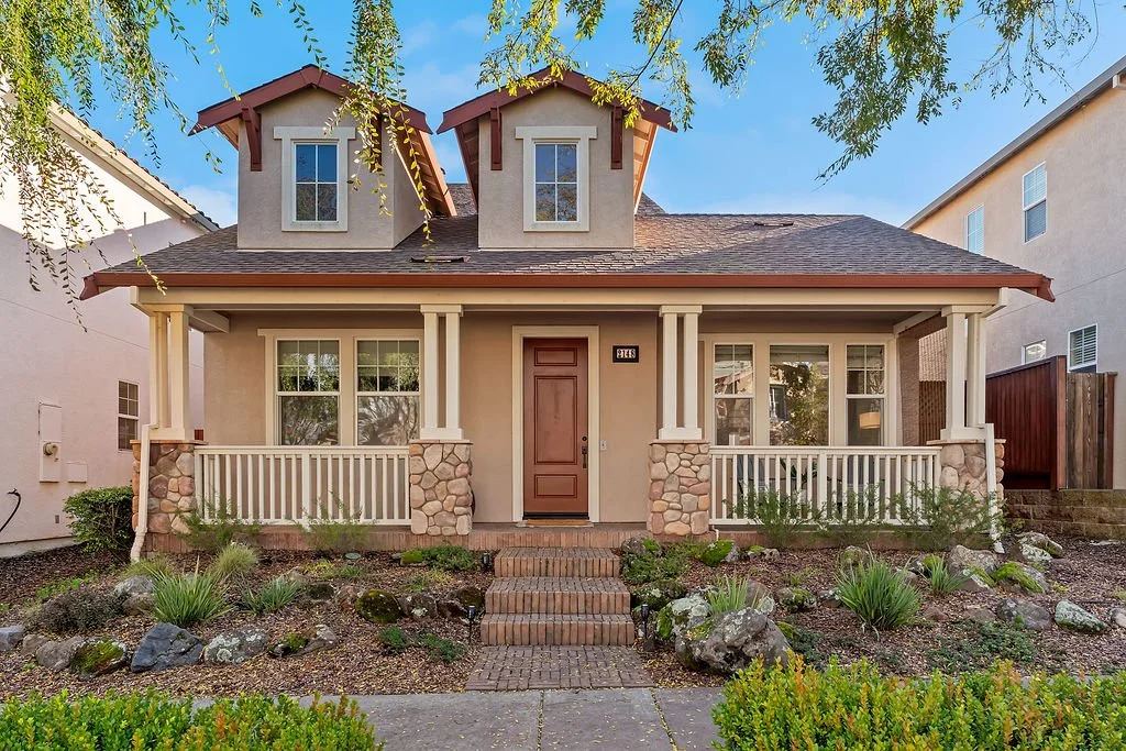 Front view of a house with a porch, stone pillars, and a wooden door. The house has a gabled roof with two dormer windows and is surrounded by a landscaped yard with plants and rocks.
