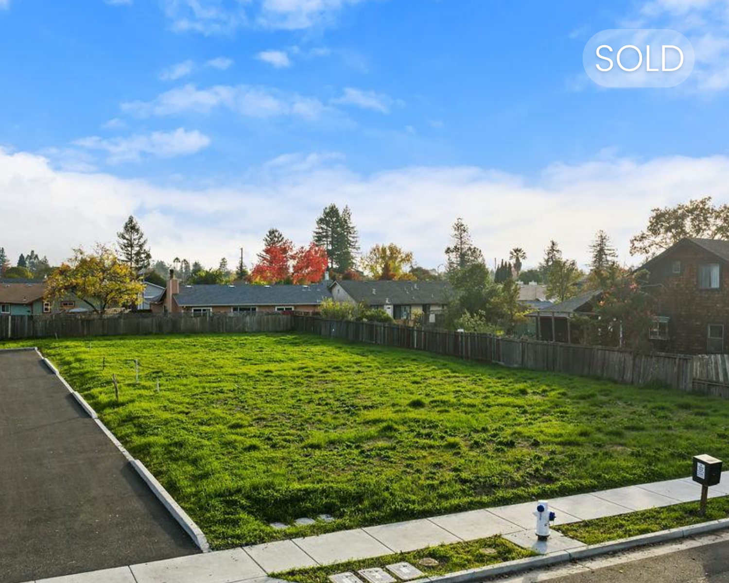 Empty suburban backyard with green grass, surrounded by a wooden fence, with neighboring houses and trees in the background under a partly cloudy sky.