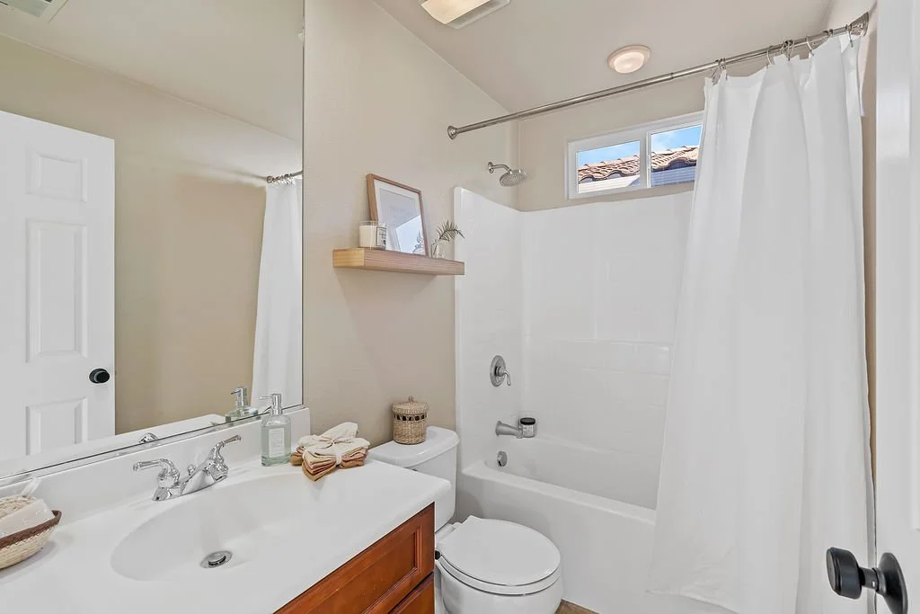 A small bathroom with a white bathtub and shower, a white toilet, a sink with a wooden cabinet, a mirror, a small window with a view of a roof, and a shelf with decorative items.
