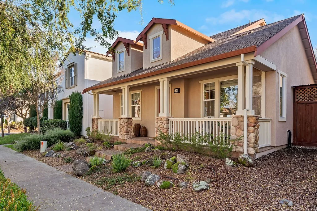 A two-story house with a porch, beige stucco exterior, stone accents on the porch columns, and a brown shingle roof. There are windows with white trim, a landscaped front yard with rocks, plants, and a sidewalk.