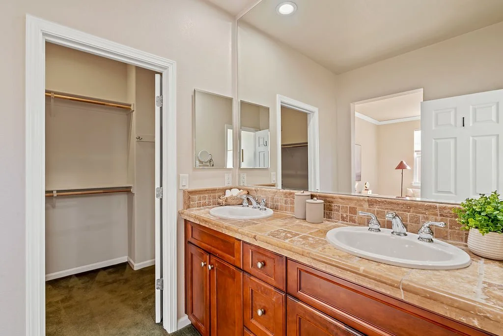 A bathroom with dual sinks, a large mirror, and a walk-in closet with open door. The countertop is beige marble, and there is a small plant on the right side of the counter.