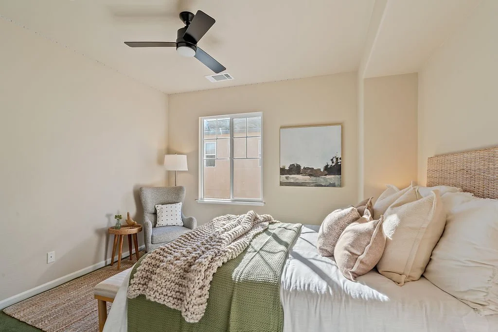 A cozy bedroom with a bed featuring cream-colored pillows and blankets, a gray armchair with a white pillow, a small wooden side table with a plant and decorative item, a window with sunlight, a framed landscape painting, beige walls, and a ceiling f