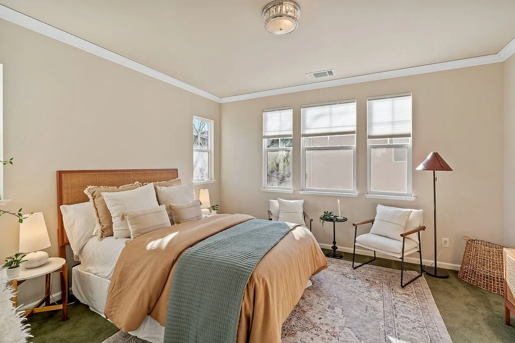 A bright, cozy bedroom with a large bed, multiple pillows, beige and white bedding, a small side table with a lamp and plants, two accent chairs, a floor lamp, and windows with white blinds.