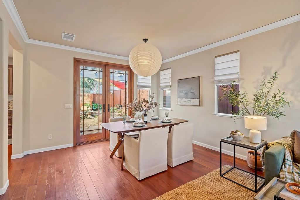 A dining room with a wooden dining table, four white slipcovered chairs, white window shades, and a large paper lantern hanging from the ceiling. There is a sliding glass door leading outside, and a small side table with a lamp and potted plant next 