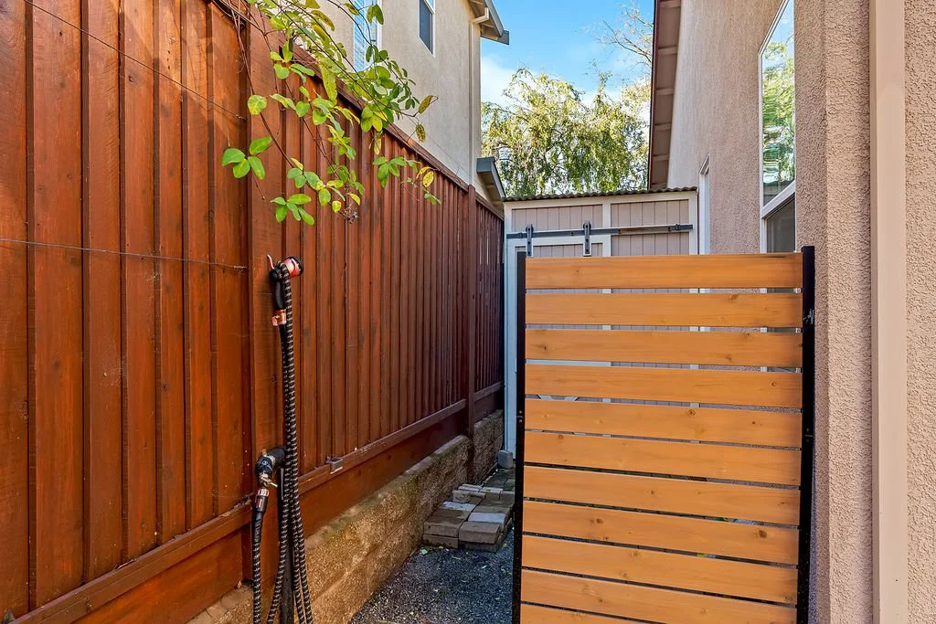 An outdoor backyard scene showing a wooden fence, a metal gate with wooden slats, a garden hose hanging on the fence, a small pathway made of brick pavers, and some greenery, with a house wall visible on the right and top background.