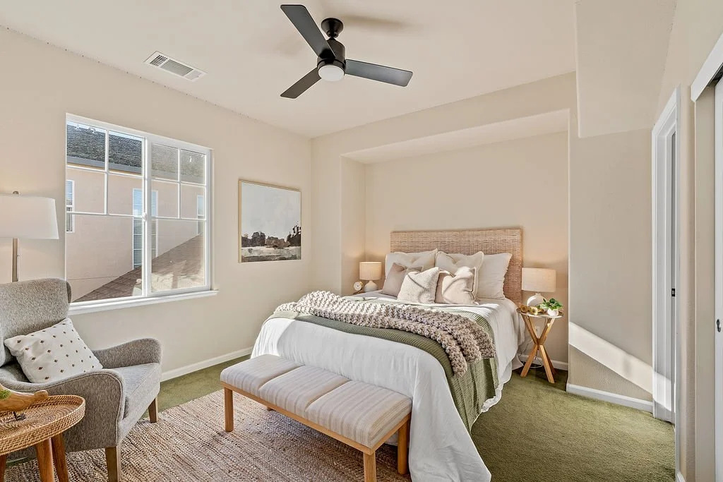 A tidy bedroom with a bed, side tables with lamps, a green and white blanket, a bench at the foot of the bed, an armchair with a pillow, a window with sunlight, a ceiling fan, a framed picture on the wall, and a closet with sliding doors.