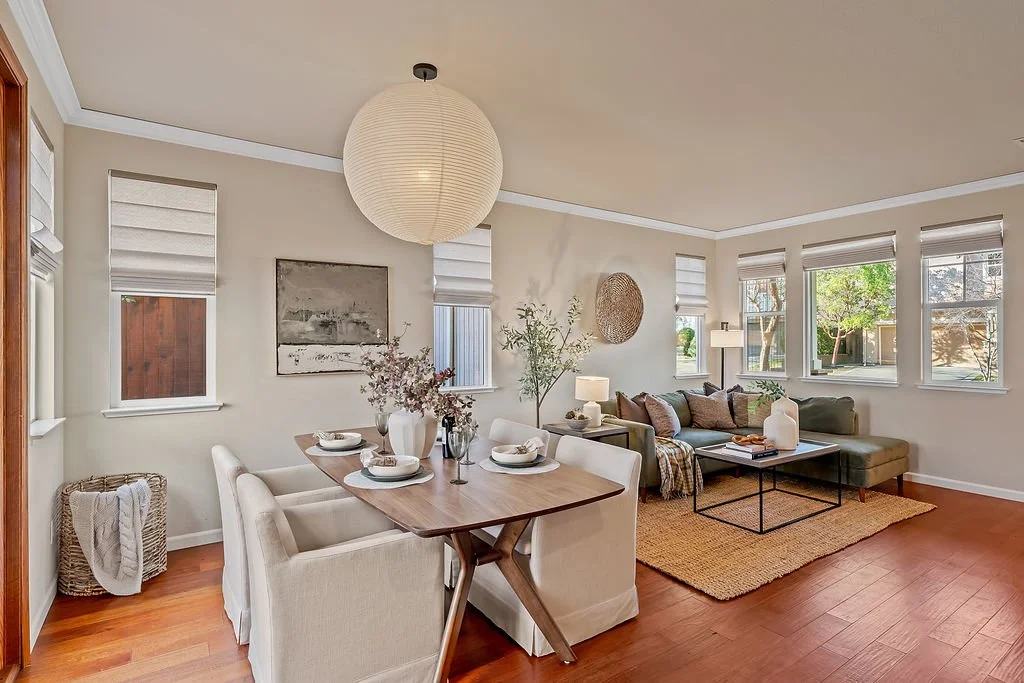 A living room and dining area with hardwood floors, beige walls, blinds on the windows, a large paper lantern ceiling light, a beige sectional sofa with throw pillows, a wooden dining table with chairs, and decorative elements like vases, wall art, a