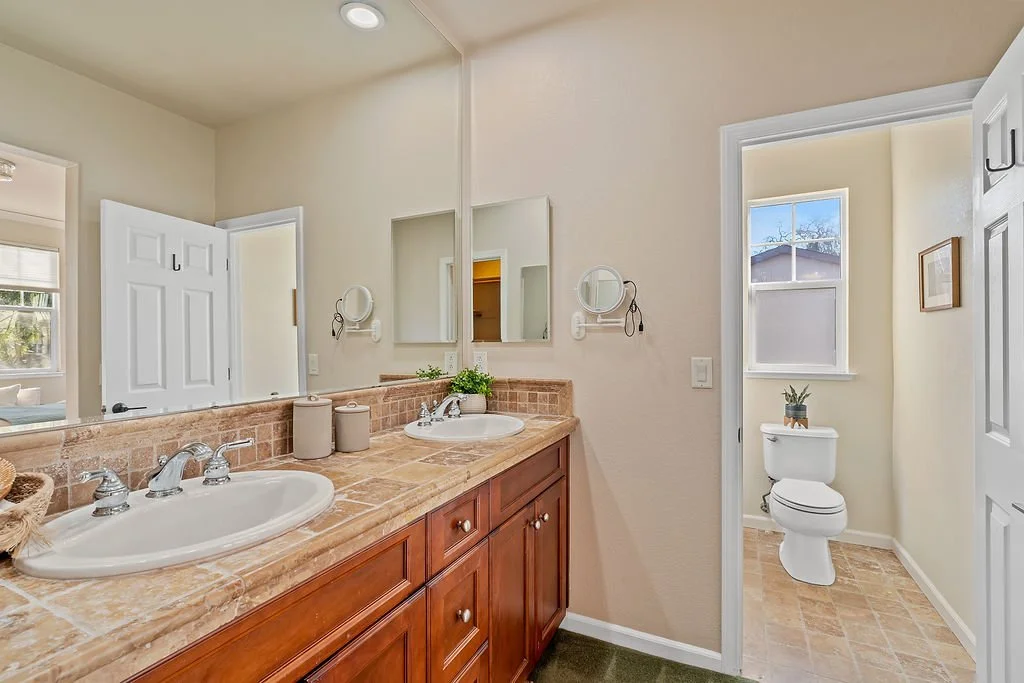 Bathroom with a dual sink vanity with brown marble countertop, wall-mounted mirrors, and a toilet in a separate room with a window and a small potted plant.