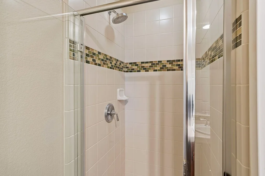 A tiled shower with a chrome showerhead, a tempered glass door, and decorative tile band on white tiles.