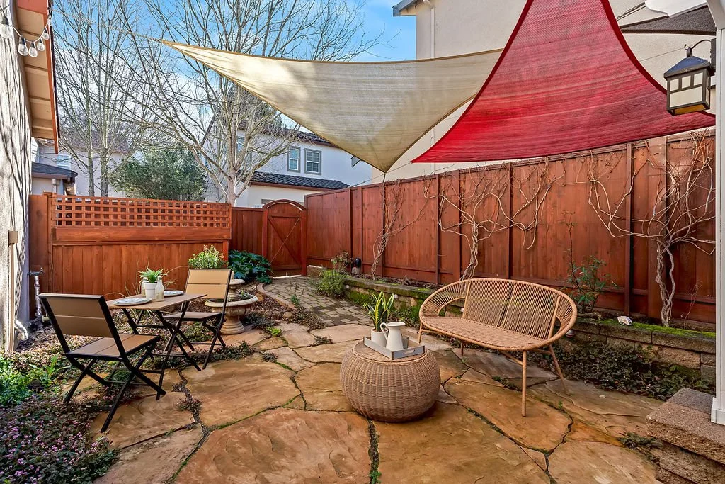 A small backyard patio with a stone floor, a brown wicker chair, a round wicker table with potted plants, a table with two black chairs, a water fountain, and a red shade sail. Wooden fence with climbing plants, trees, and neighboring houses in the b