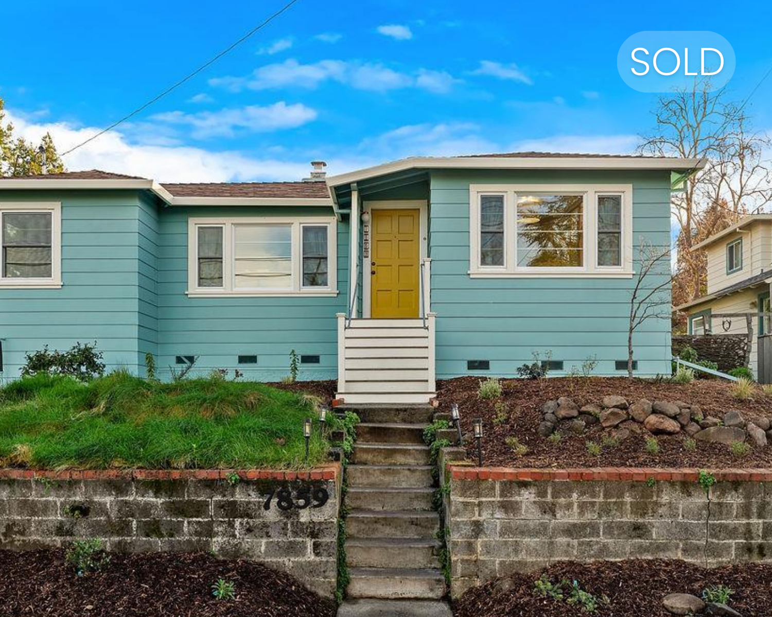 Front view of a light blue house with a yellow door, white stairs, and a sold sign in the upper right corner.