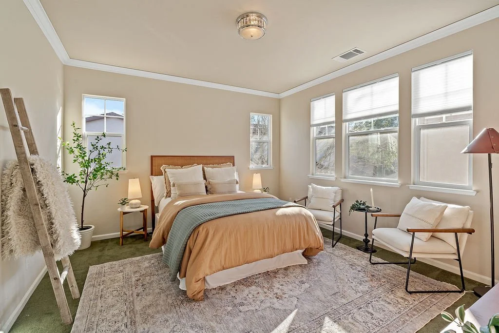 Bright bedroom with beige walls, a bed with beige and blue bedding, wooden headboard, two side tables with lamps, two white chairs, windows with blinds, a potted plant, and a decorative ladder with a fur throw.