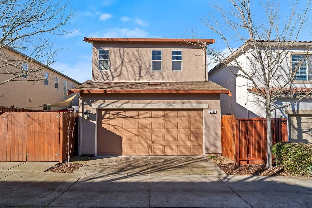A two-story modern house with a brown garage door, surrounded by wooden fences and leafless trees, under a clear blue sky.