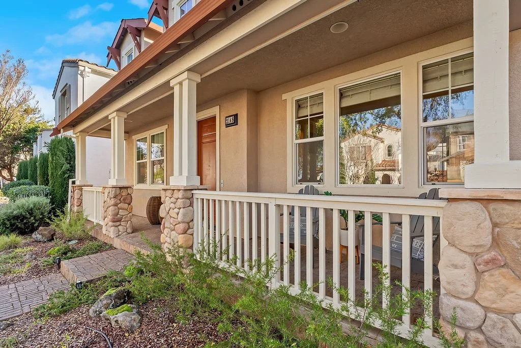 Front porch of a house with stone pillars, white railing, and outdoor seating, surrounded by landscaping and neighboring homes.