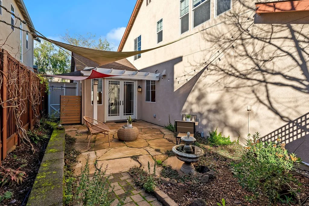Small backyard patio with stone paving, a fountain, potted plants, wicker chair, and wall decorations, enclosed by a fence and neighboring buildings, with sun and shadows cast by trees.