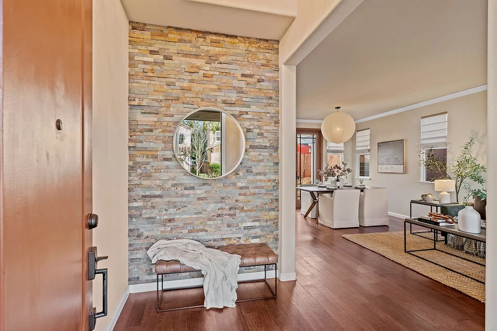 Entryway with a stone accent wall, a round mirror, a brown bench with a blanket, and a view into a dining and living area with white walls, wooden floors, and modern decor.