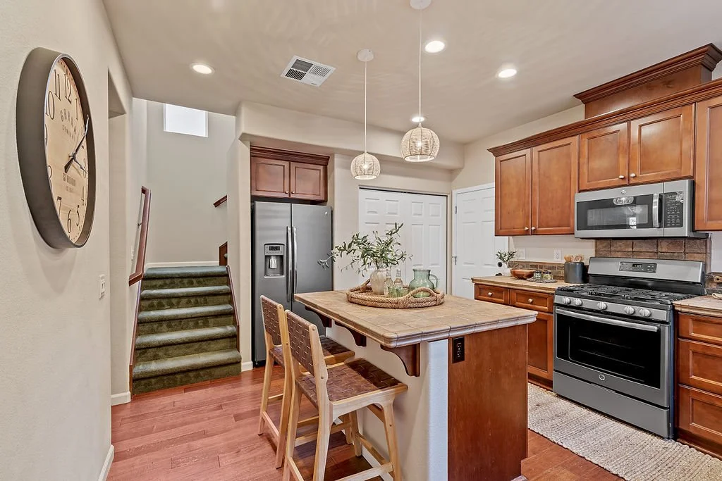 A modern kitchen with wooden cabinets, a stainless steel refrigerator, a black stove, microwave, and a kitchen island with two chairs. There is a large wall clock to the left and a staircase leading upstairs in the background.