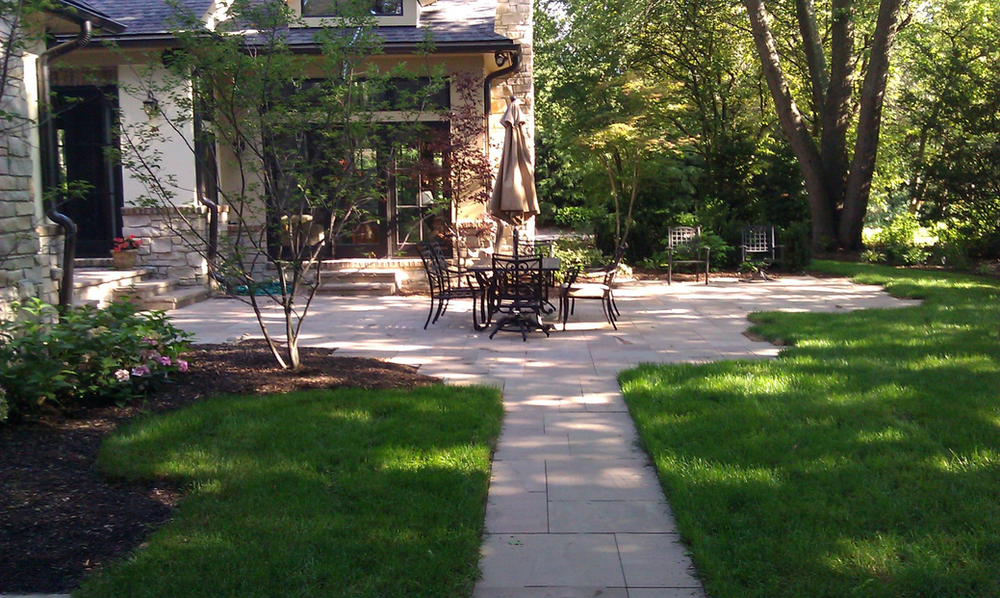 Backyard patio with stone tile flooring, outdoor dining table with chairs and umbrella, surrounded by trees and greenery, and steps leading to a house.