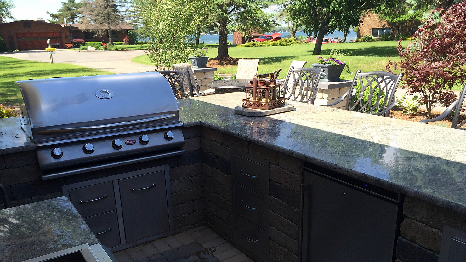 Outdoor kitchen with a stainless steel grill, granite countertops, and patio chairs on a sunny day in a green backyard.