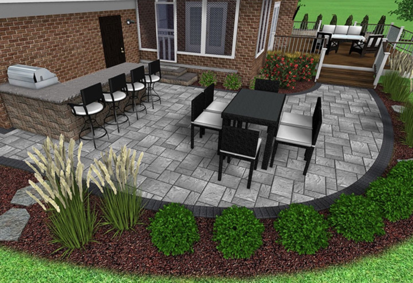 A backyard patio with a brick house in the background, featuring a raised deck with outdoor furniture, a lower paved area with a dining table and chairs, and decorative plants and bushes surrounding the space.
