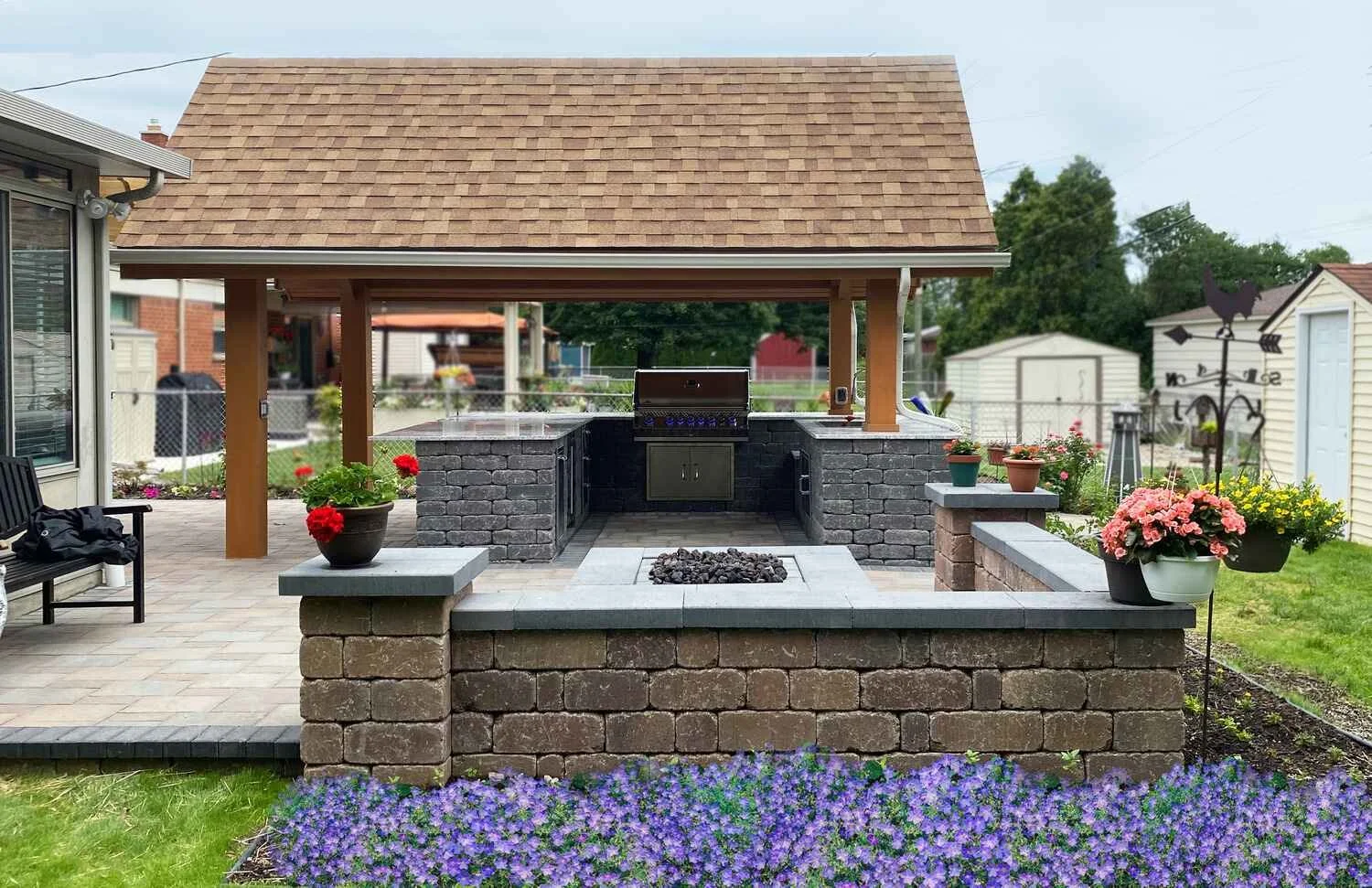 Backyard outdoor kitchen with grill, brick and stone counter, in a fenced yard with colorful flowers and garden decorations.