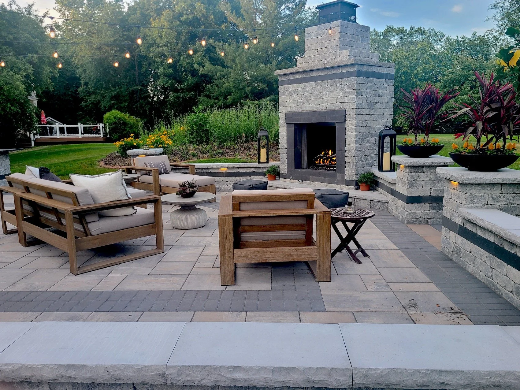 Outdoor patio with stone flooring, wooden furniture with cushions, a central firepit, plants, and a brick fireplace with a fire, surrounded by string lights and a garden with trees in the background.